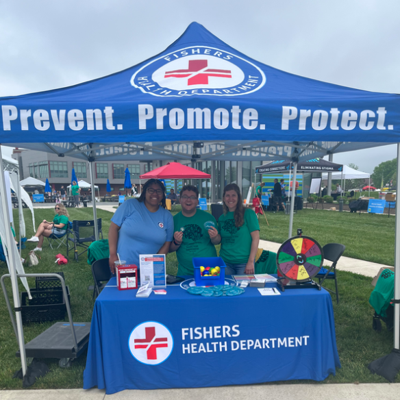 Three volunteers stand at a blue Fisher's Health Department tent with 'Prevent. Promote. Protect.' on the canopy, a wheel game and health‑info materials on the table.]