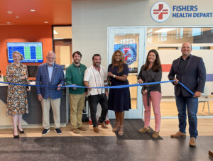 Seven people stand behind a blue ribbon during a ribbon-cutting at a health department, with a Fishers Health Department sign visible.