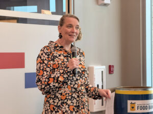 Woman speaking into a handheld microphone at a community event, wearing a orange–black floral dress, beside a food bank donation barrel.