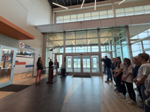 Speaker at podium addressing a line of people in a lobby with Fishers Health Department signage nearby and a camera crew recording.