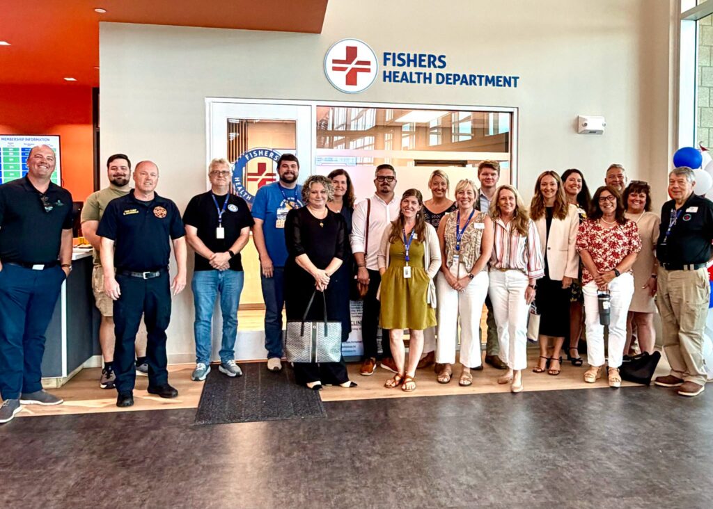 Group of staff and visitors posing for a photo in the Fisher's Health Department lobby, with the department sign and logo behind them.