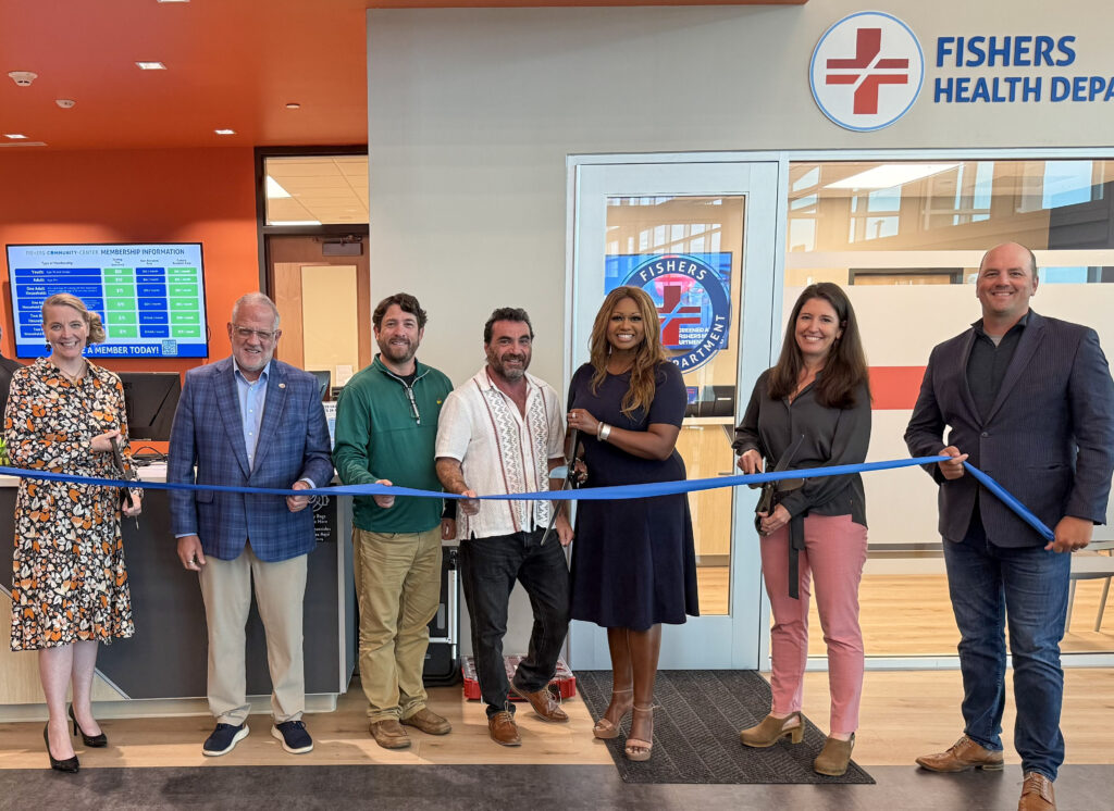 Seven people stand behind a blue ribbon during a ribbon-cutting at a health department, with a Fishers Health Department sign visible.