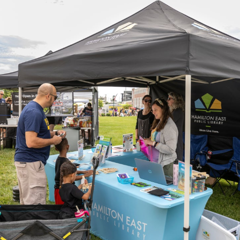 Outdoor festival scene with a Hamilton East Public Library booth under a black canopy; staff assist a man and two children at a table with brochures and a laptop.