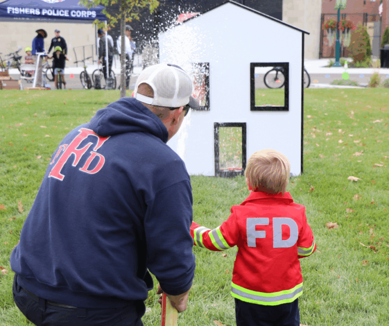 A firefighter and young child in FD jackets hold hands in a park during a community safety event, with a white playhouse prop and water spray in the background.